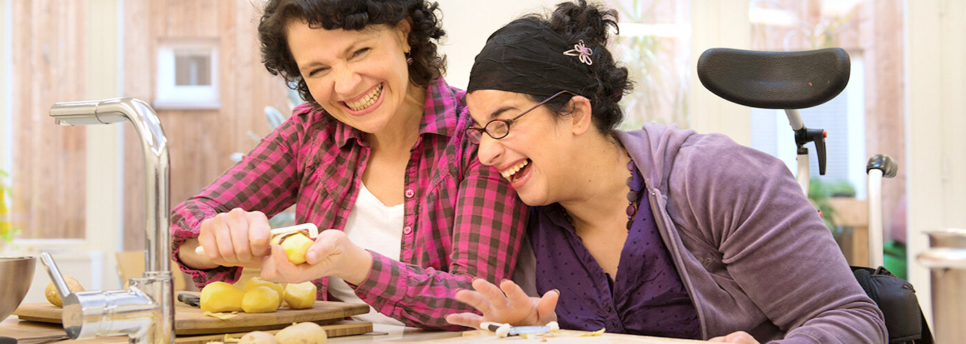 Eine Betreuerin sitzt gemeinsam mit einem geistig und körperlich behinderten Mädchen an einem Tisch in der Küche. Beide schälen Kartoffeln und lachen dabei herzlich.