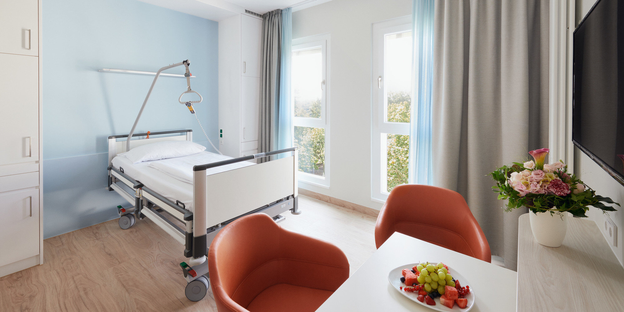 Blick in ein modern eingrichtetes Komfortzimmer des Martin Luther Krankenhauses mit einem Tisch, zwei Stühlen, einem Pflegebett sowie Dekoration.