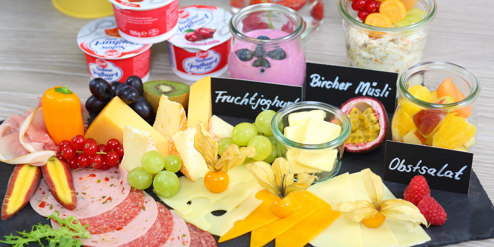 Auf einem Schieferbrett angerichtete Wurst- und Käseplatte mit Obst und essbarer Dekoration. Im Hintergrund befinden sich Joghurtbecher, Dessertgläser und Getränke.