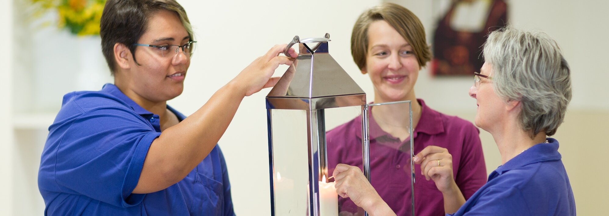 Drei Mitarbeiterinnen entzünden gemeinsam eine Kerze in einer Edelstahllaterne mit Glasfenstern