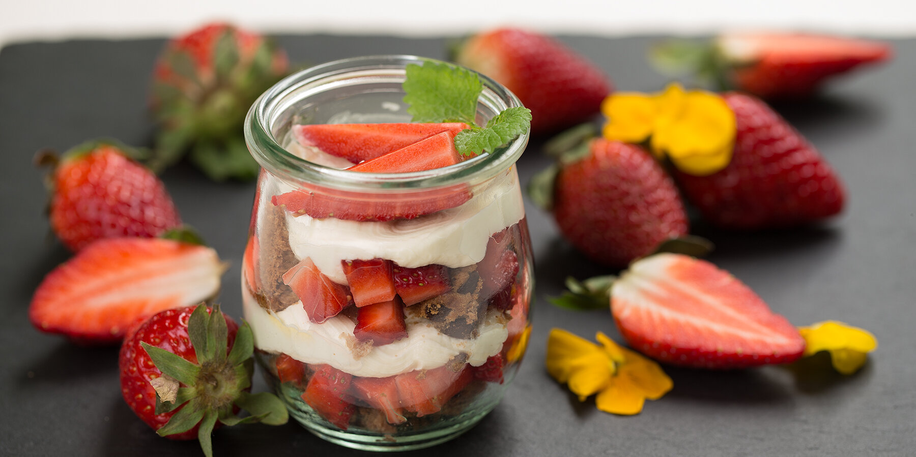 Ein Schieferbrett auf dem ein Glas mit Dessert aus Erdbeeren und Milchcréme steht. Um das Glas herum sind Erdbeeren und Blütenblätter drapiert.