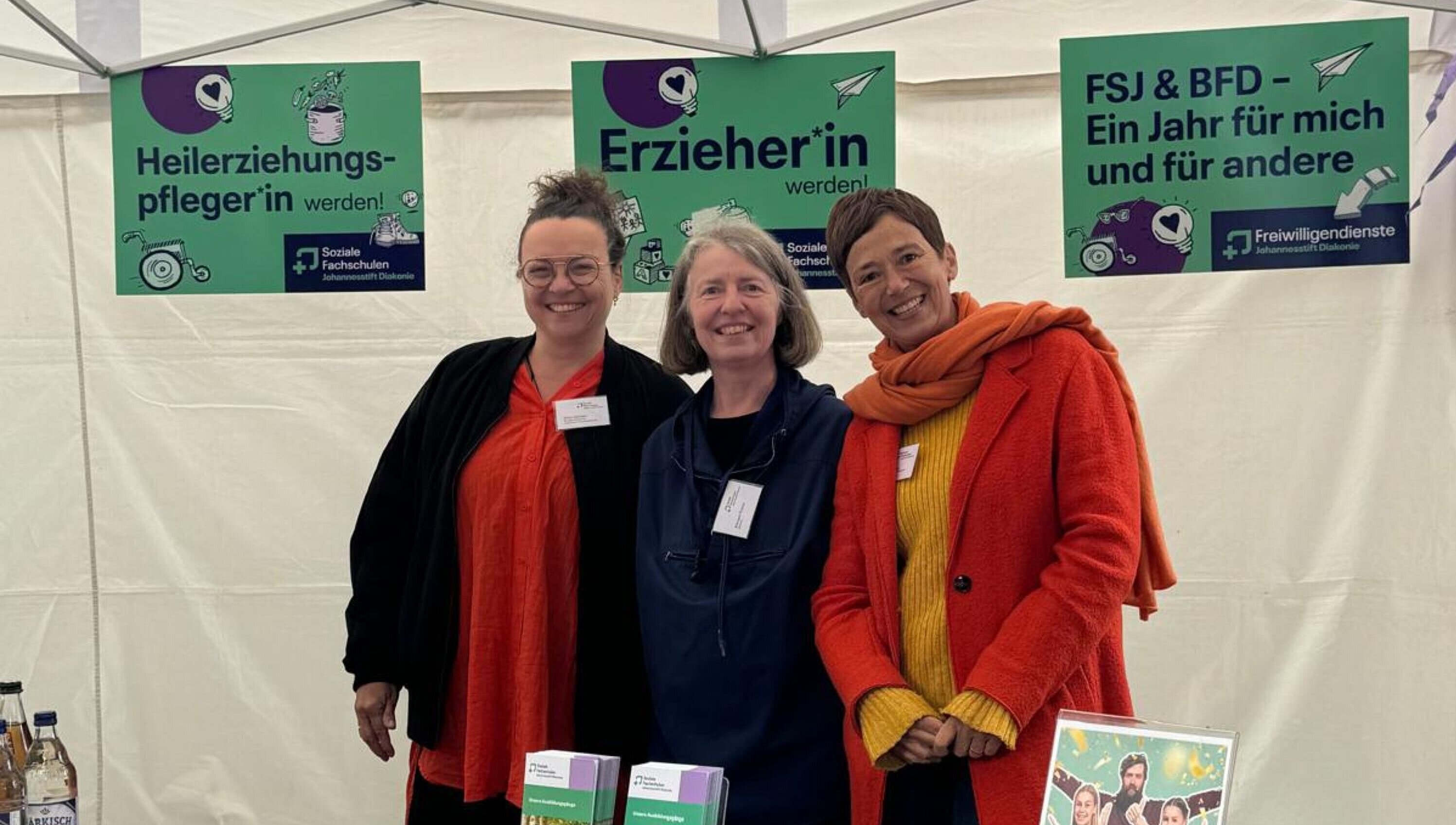 Drei Mitarbeitende stehen an einem Stand, hinter ihnen bunte Schilder.