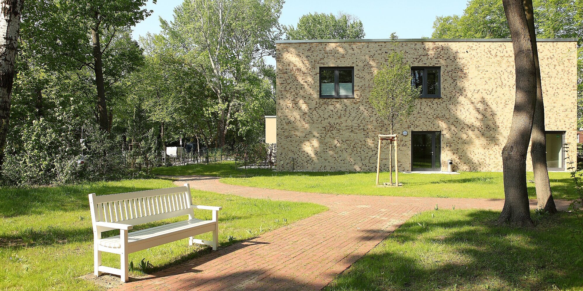 Eine weiße Bank, mehrere Bäume und ein angelegter Weg im Hospizgarten, im Hintergrund das Paul Gerhardt Hospiz.