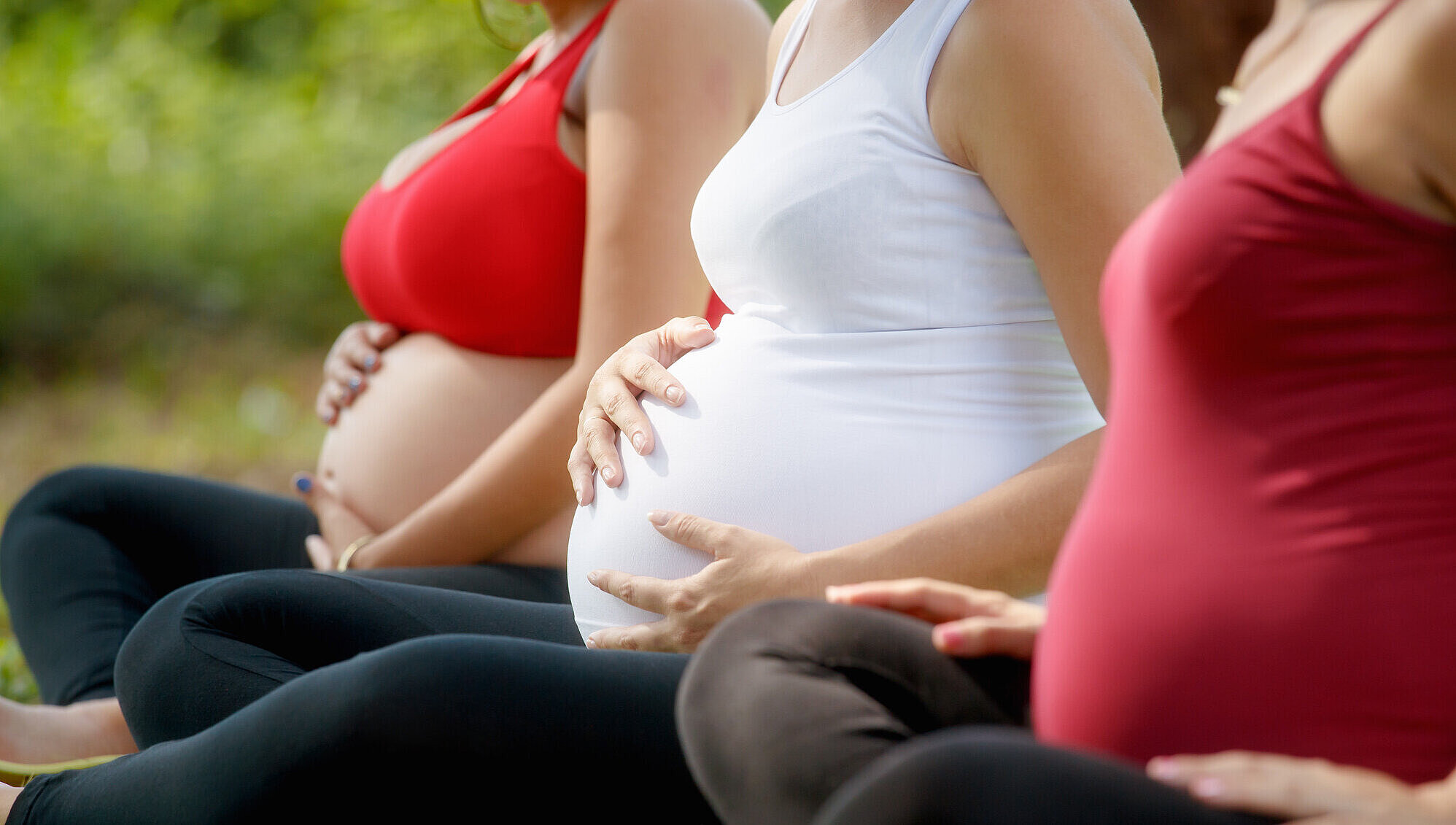 Drei schwangere Frauen sitzen im Park. Zwei davon halten sich den Babybauch.