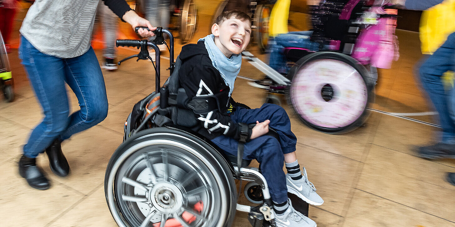 Wöchentliches Treffen für tanzbegeisterte Schüler*innen Kinder, größtenteils im Rollstuhl, und mehrere Betreuer*innen bewegen sich tanzend im Kreis. In der Mitte ist ein Junge im Rollstuhl zu sehen.