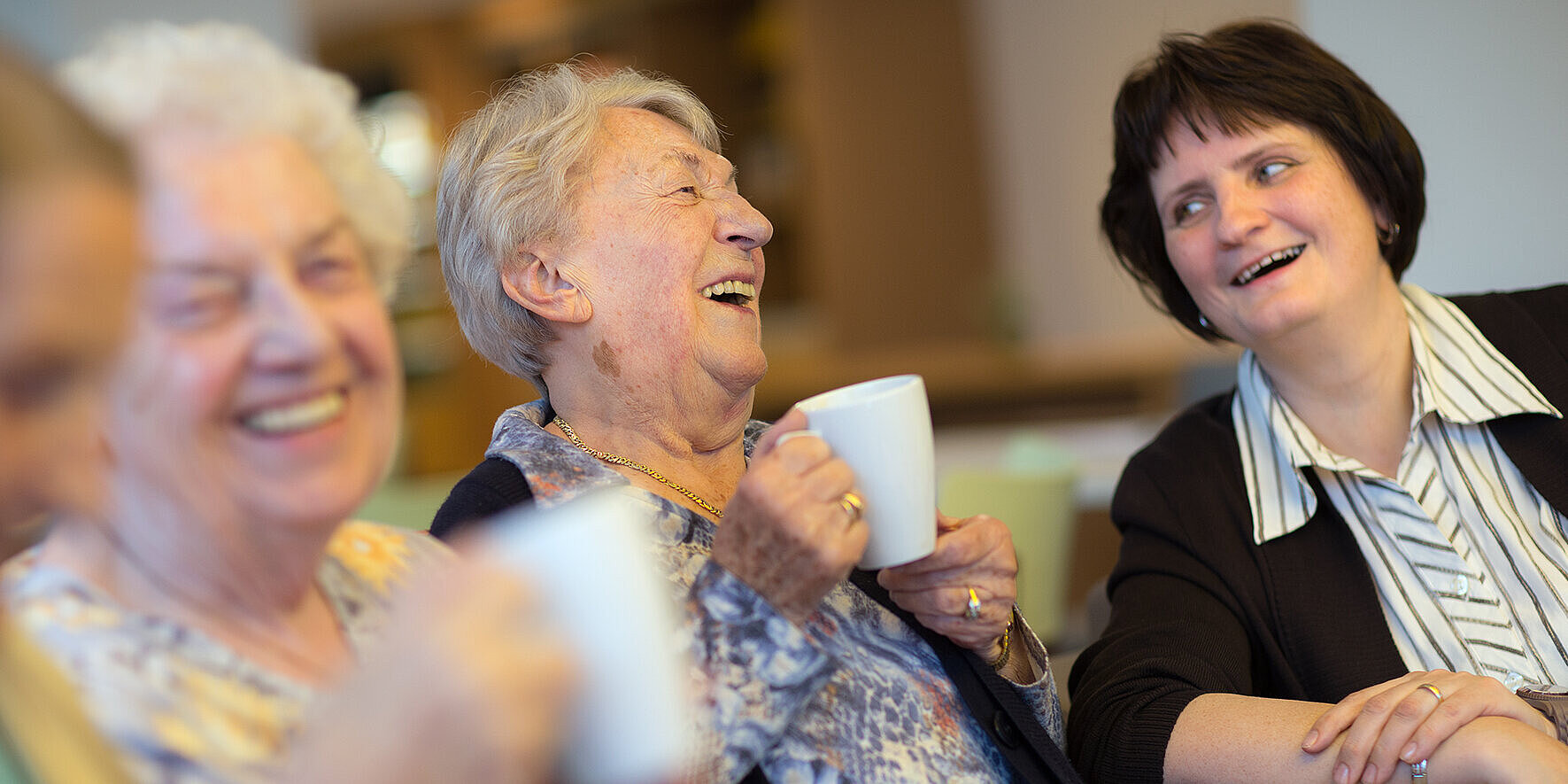 Bewohnerinnen und Angehörige trinken gemeinsam Kaffee und lachen sie sich gegenseitig an