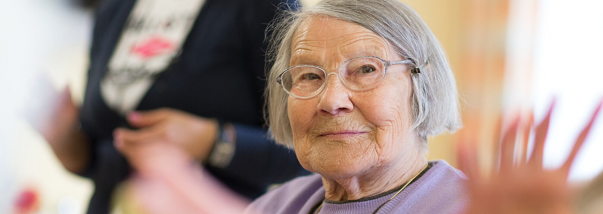 Seniorin bei Kaffee und Kuchen, im Hintergrund steht eine Pflegekraft.