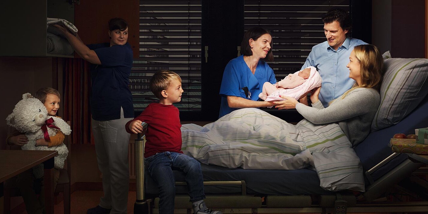Eine Hebamme reicht einer im Krankenhausbett liegenden Frau ihr neugeborenes Kind. Neben der Frau steht der Vater, am Fußende des Bettes sind zwei Söhne zu sehen. Im Hintergrund räumt eine Pflegerin Wäsche in einen Schrank. Alle Personen schauen lächelnd das Baby an.