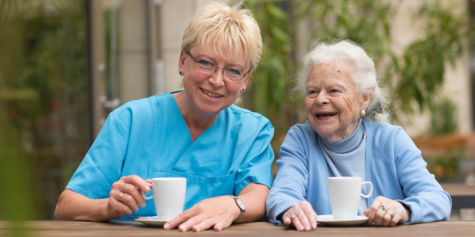 Eine Pflegekraft sitzt mit einer älteren Dame im Garten. Beide halten eine Kaffeetasse in der Hand und lachen.