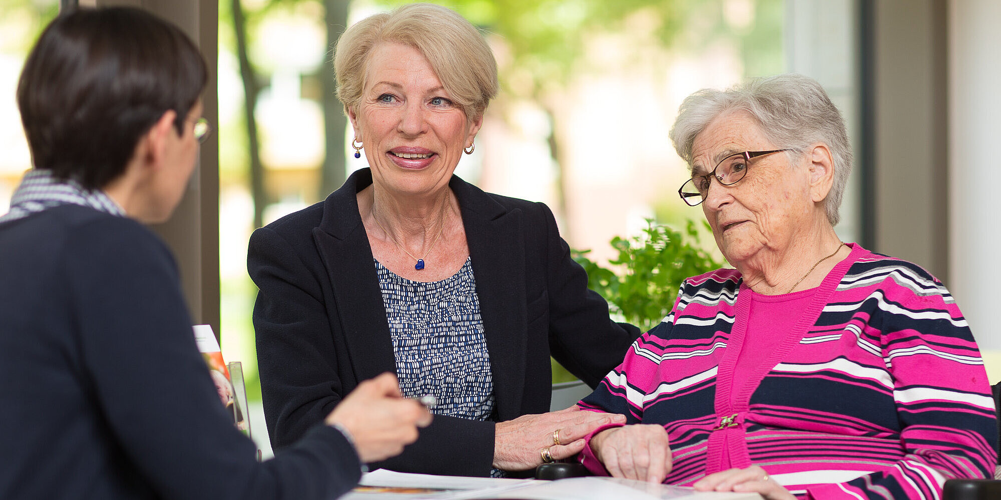 Mitarbeiterin des Hauses berät ältere Frau und deren Tochter. Auf dem Tisch liegt eine Informationsbroschüre.