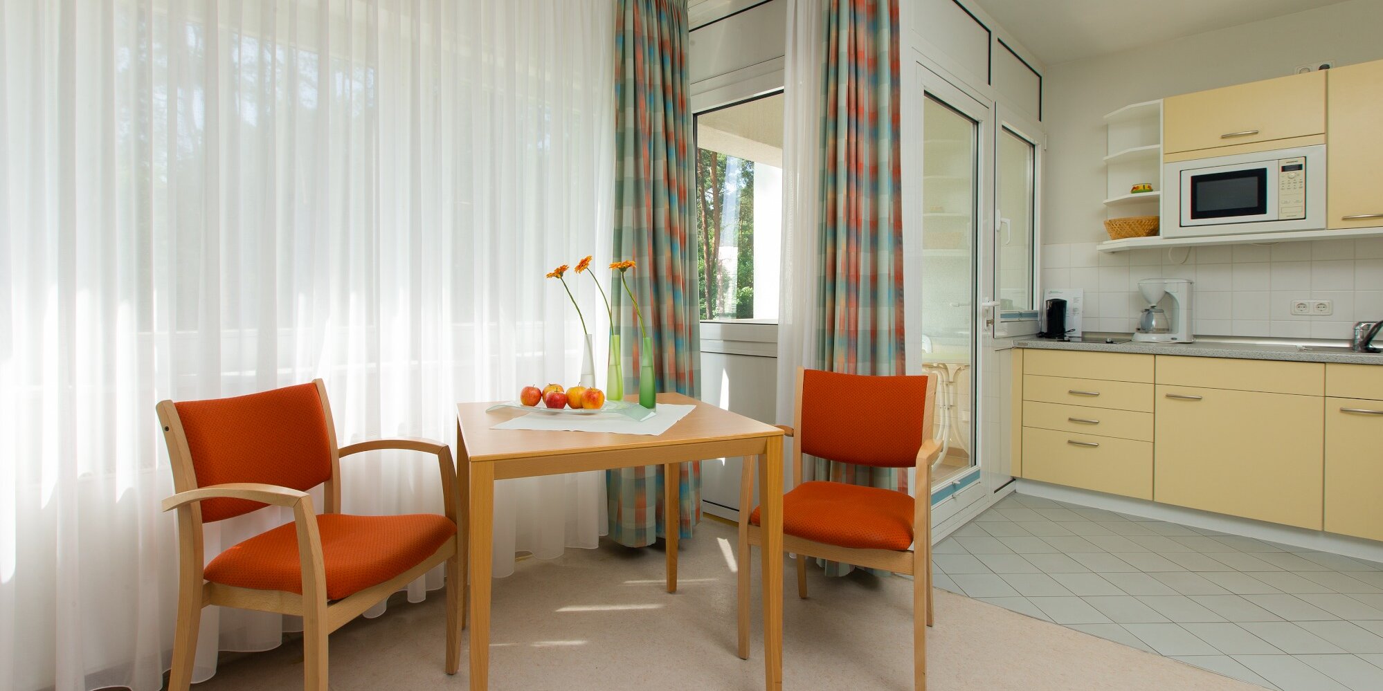 Modern eingerichtetes Appartement mit einer Sitzecke, bestehend aus einem kleinen Tisch und zwei Stühlen. Eine kleine komplett ausgestattete Küchenzeile schließt direkt an den Balkon an.