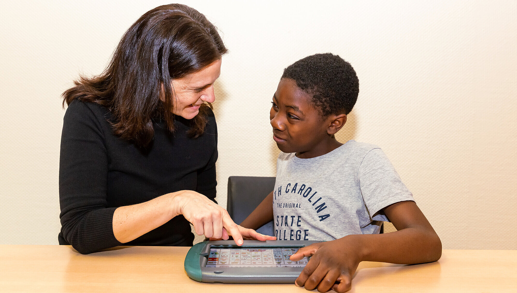 Lernen mit elektronischen Kommunikationshilfen Ein Schüler und seine Lehrerin sitzen mit einem Tablet am Tisch und schauen einander an.