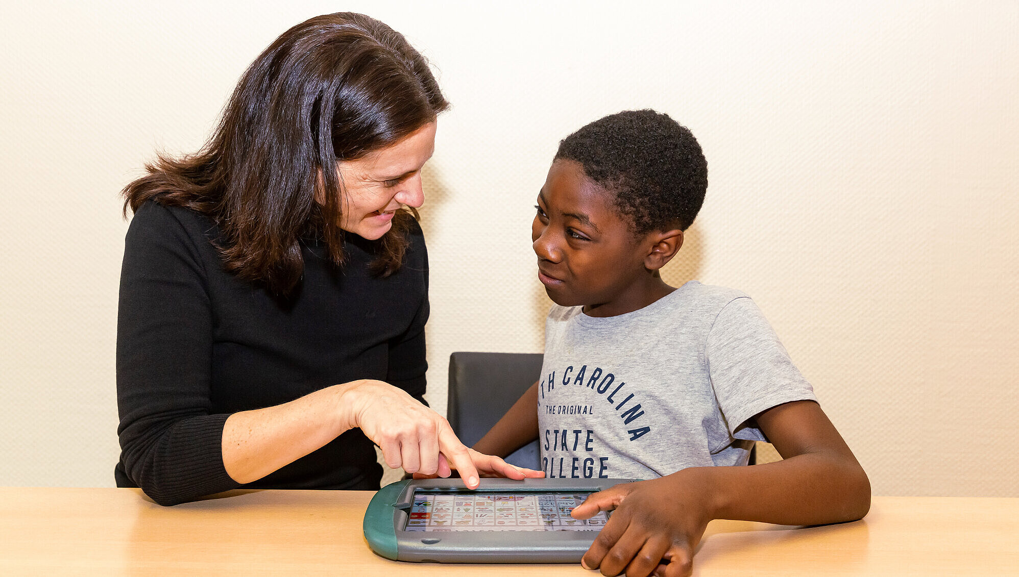 Lernen mit elektronischen Kommunikationshilfen Ein Schüler und seine Lehrerin sitzen mit einem Tablet am Tisch und schauen einander an.
