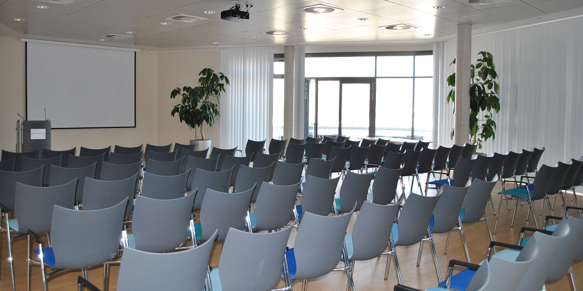 Ein Saal mit Reihenbestuhlung. Vorne befindet sich ein Rednerpult und eine Leinwand sowie unter der Decke ein Beamer.