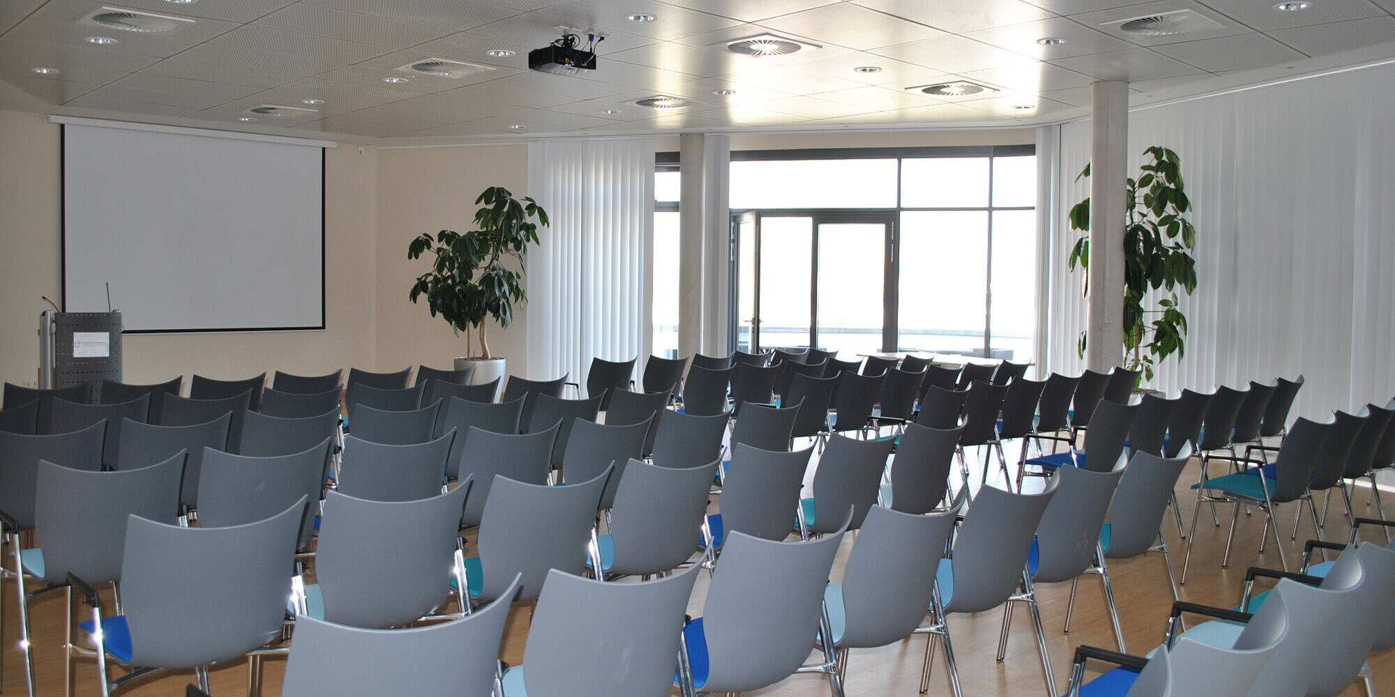 Ein Saal mit Reihenbestuhlung. Vorne befindet sich ein Rednerpult und eine Leinwand sowie unter der Decke ein Beamer.
