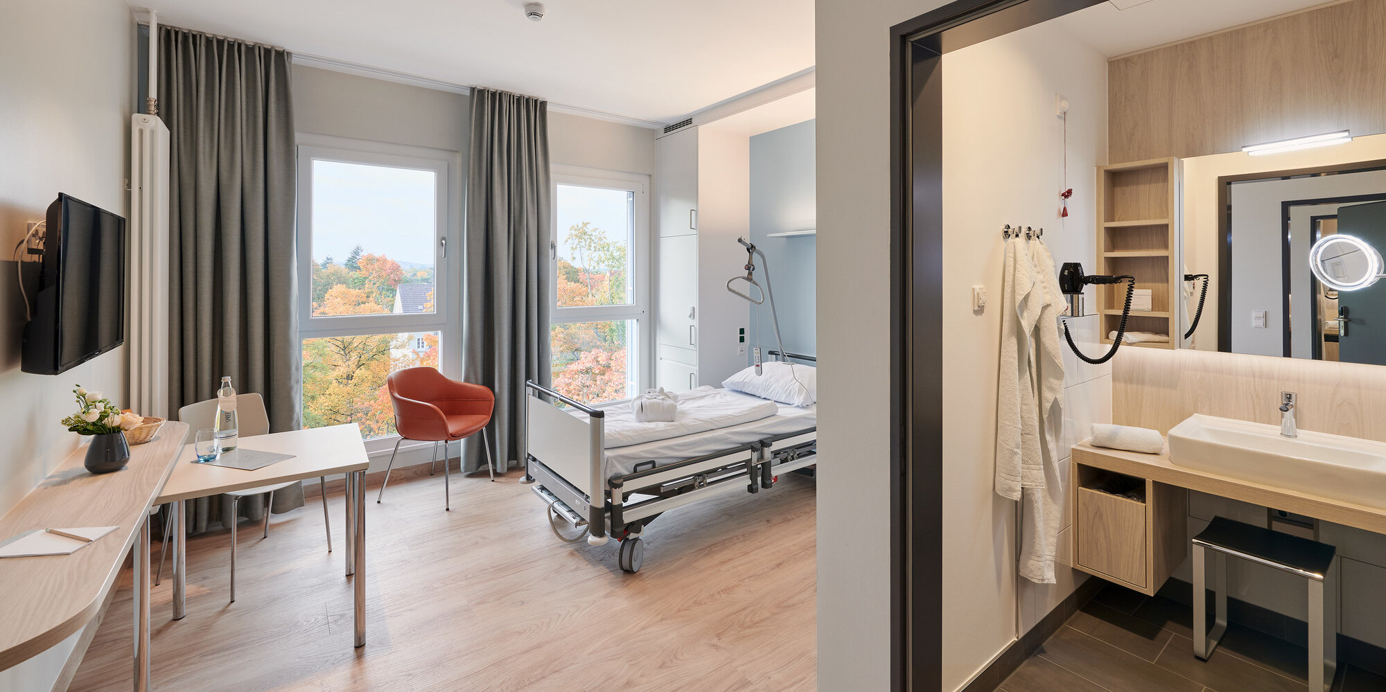 Blick in ein modern eingerichtetes Komfortzimmer des Martin Luther Krankenhauses mit eigenem Bad, Schreibtisch, Pflegebett, Sitzgelegenheit und Flachbildschirm an der Wand.