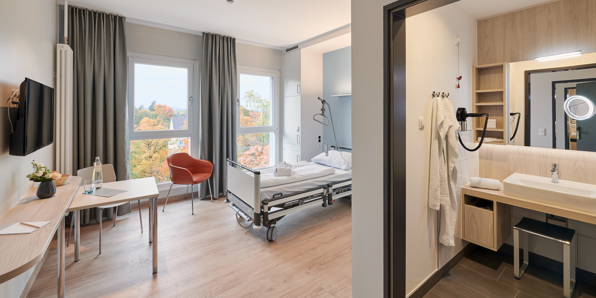 Blick in ein modern eingerichtetes Komfortzimmer des Martin Luther Krankenhauses mit eigenem Bad, Schreibtisch, Pflegebett, Sitzgelegenheit und Flachbildschirm an der Wand.