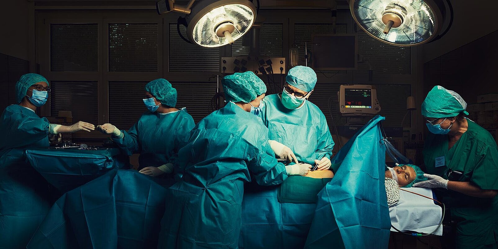 Ein Patient liegt auf dem OP-Tisch in einem modernen Operationssaal. In der Bildmitte operiert ein Arzt. Dabei spricht er mit einer Pflegekraft, die ihm assistiert. Rechts hält eine Anästhesistin den Kopf des Patienten fest. Im Hintergrund reicht eine weitere Pflegekraft einer Kollegin chirurgisches Instrument, dabei blicken sie sich an. Alle Personen tragen Mundschutz und türkisfarbene Schutzkleidung.