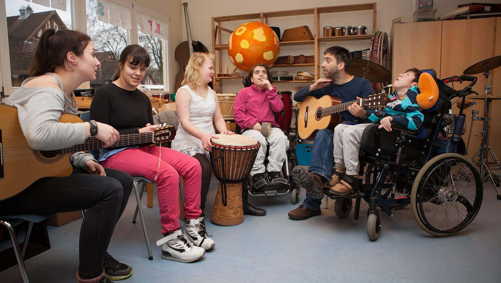 August Hermann Francke Schule: Gemeinsames Musizieren In einem Musikzimmer musizieren eine Pädagogin und ein Pädagoge mit Gitarren gemeinsam mit vier körperlich und geistig behinderten Schülerinnen und Schülern. Zwei der Schülerinnen haben eigene Instrumente, eine Trommel und eine Cabasa, mit denen sie am Unterricht teilnehmen.