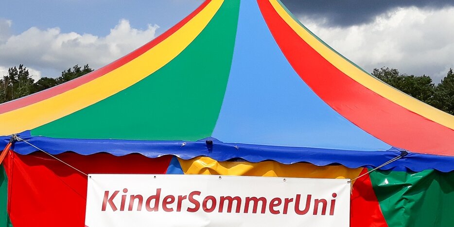 Ein bunt gestreiftes Zelt mit einem Banner der KinderSommerUni.
