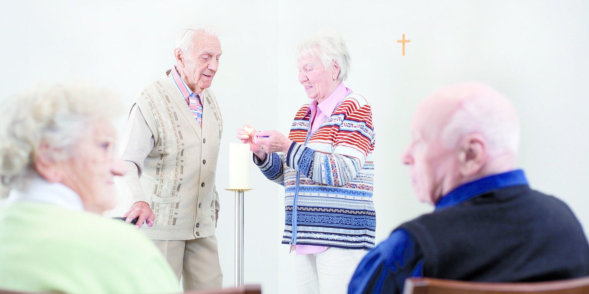 Bewohnerin und Bewohner entzünden im Hintergrund gemeinsam eine Kerze in der Kapelle. Im Vordergrund unterhalten sich eine Bewohnerin und ein Bewohner.