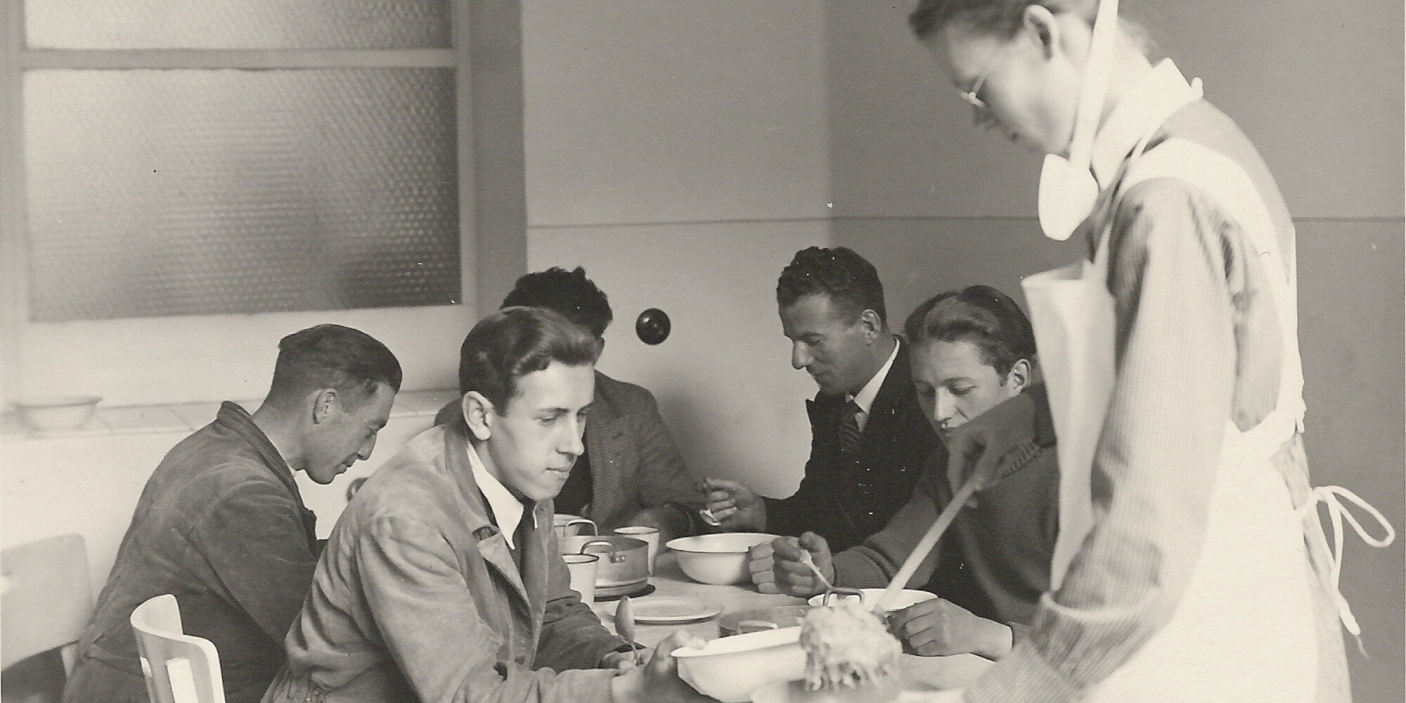 Altes schwarz-weiß-Foto: Mehrere Männer in Uniform sitzen an einem Tisch und erhalten Mittagessen von einer Pflegekraft.