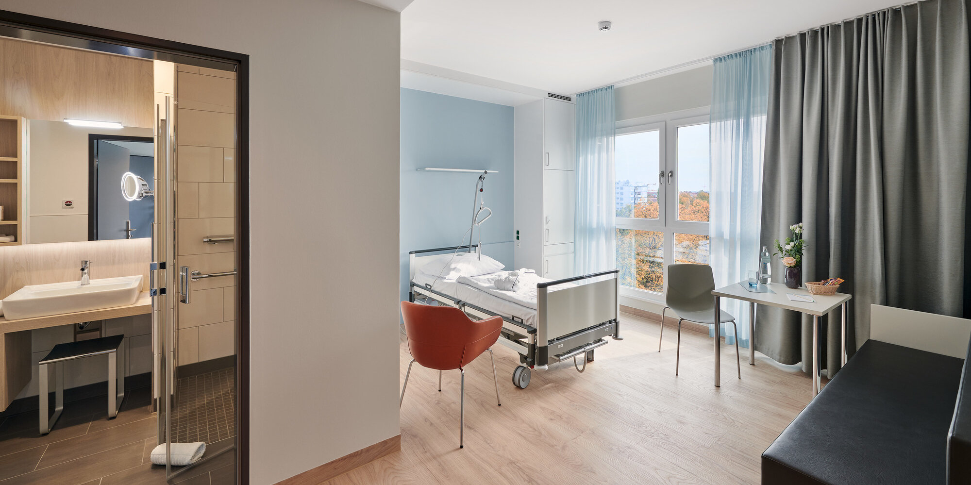 Ein Einbettzimmer mit modernem Bad und einem wohnlich eingerichteten Zimmer mit Krankenhausbett, Schreibtisch und Sitzgelegenheit für Patient*innen auf der Komfortstation.