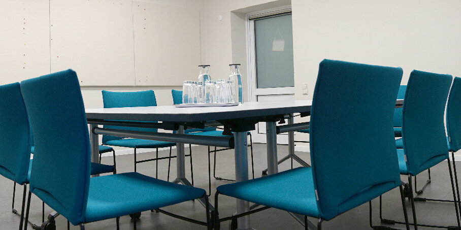 Ein heller Raum mit einem eckigen Tisch und insgesamt 12 Sitzmöglichkeiten, bestehend aus türkisfarbenen Stühlen. Der Tisch ist eingedeckt mit zwei Glaskaraffen und diversen Trinkgläsern.