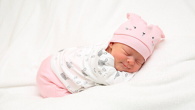 Neugeborenes Baby mit rosafarbener Mütze schläft auf einer weißen Decke.