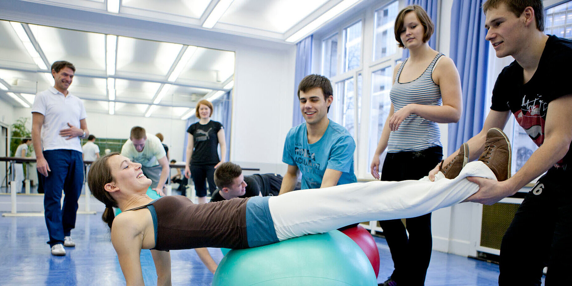 Immer in Bewegung: In unseren hochwertig ausgestatteten Übungsräumen kannst du gelernte Therapiemethoden direkt praktisch umsetzen. Schülerin liegt auf einem grünen Gymnastikball und wird von einem anderen Schüler gestützt.