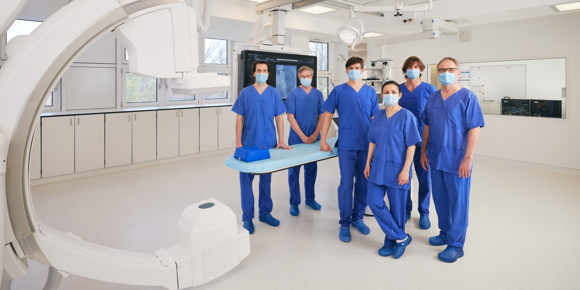 Das Team der Klinik für Kardiologie posiert in Arbeitskleidung für ein Gruppenfoto im Herzkatheterlabor.