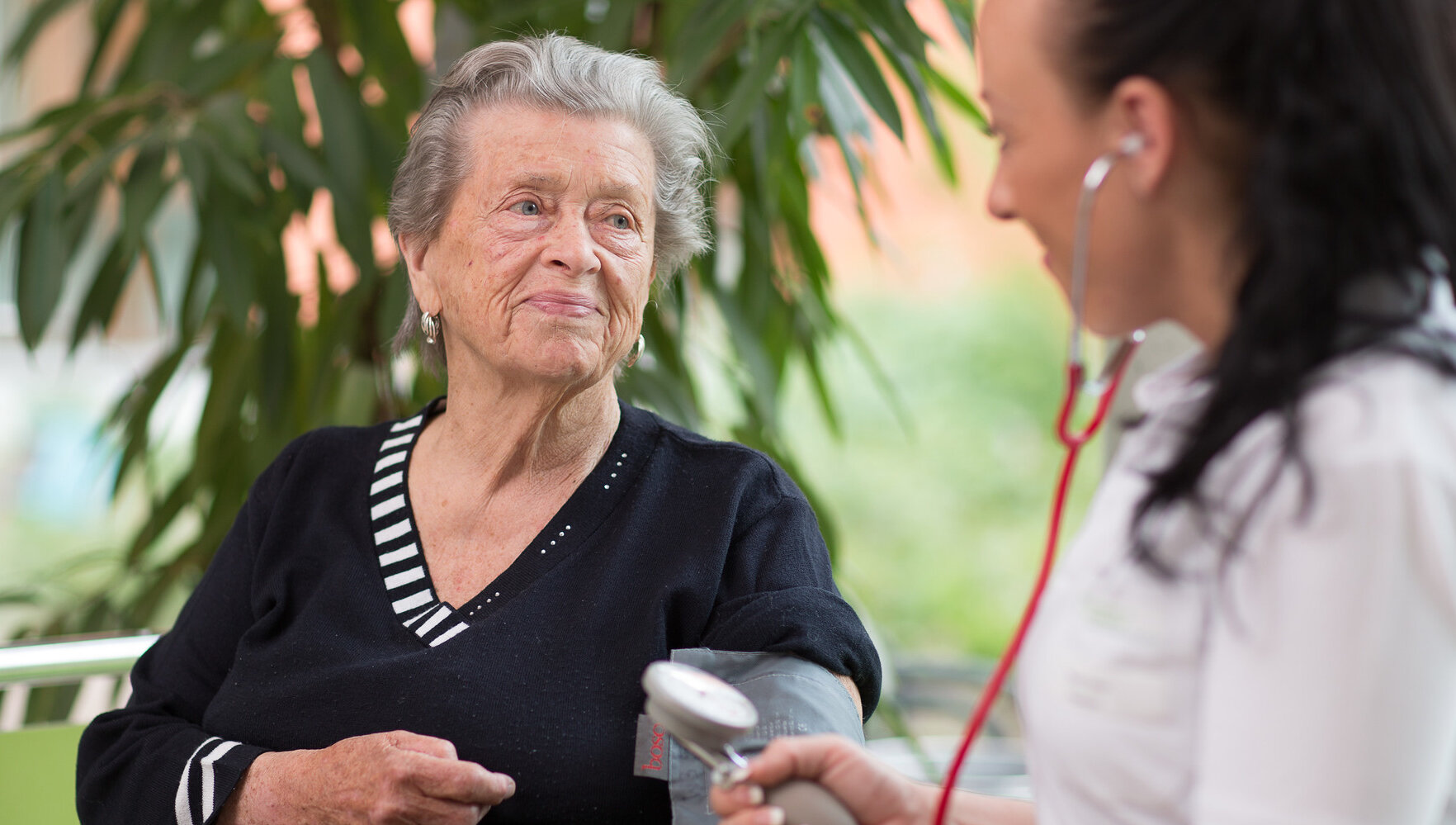 Eine Pflegekraft misst bei einer alten Frau per Manschette den Blutdruck. Beide lächeln sich dabei an.