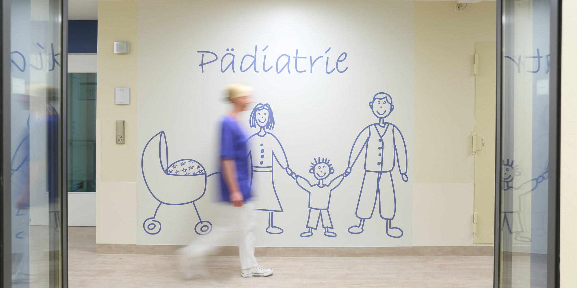 Pflegerin vor einer Wand mit der Aufschrift "Pädiatrie" und gezeichneter Familie.