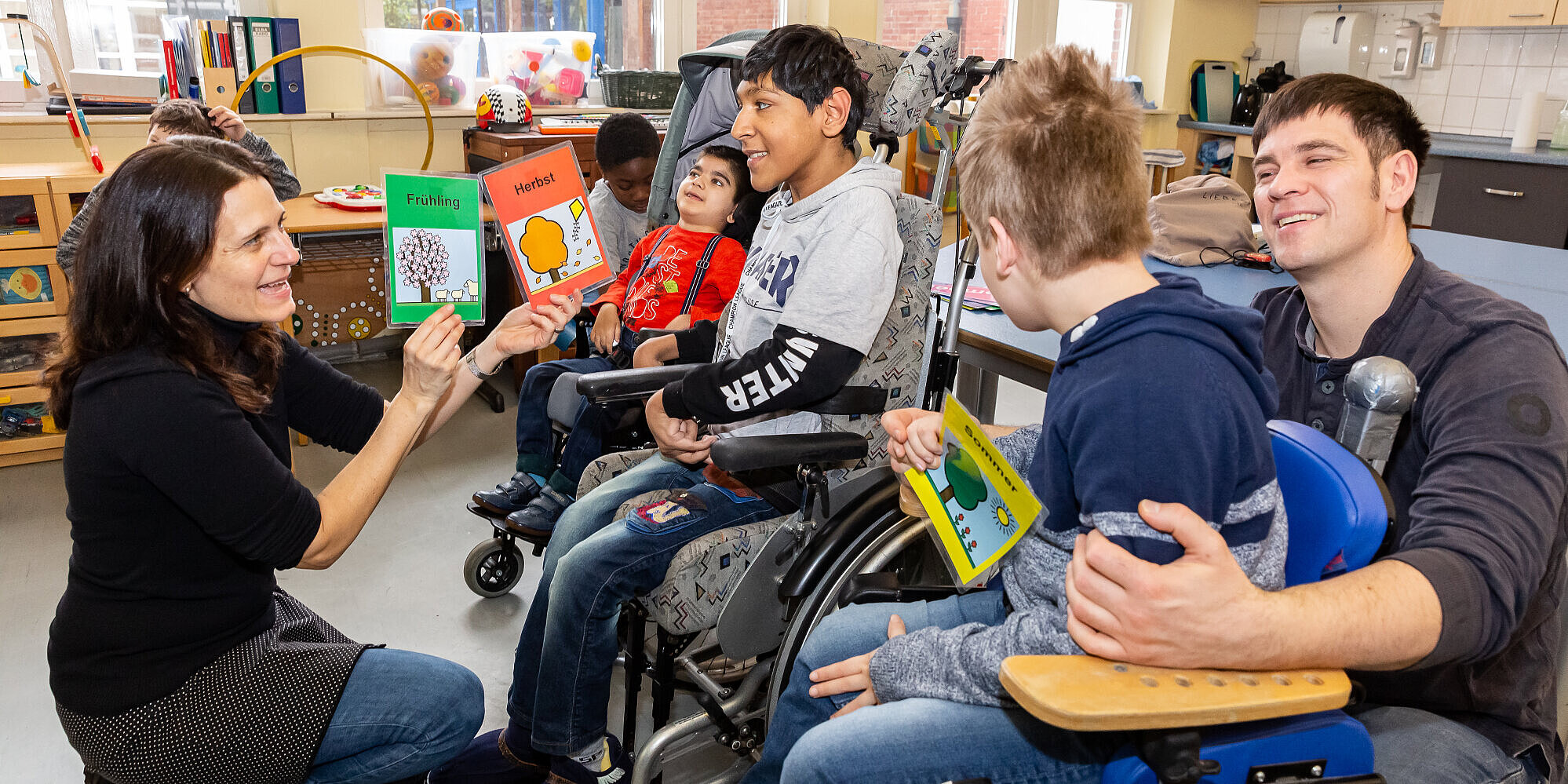 Wohlfühlatmosphäre: Kleine Klassen und eine angenehme Lernumgebung Ein Schüler und eine Lehrerin sitzen auf dem Fußboden; im Hintergrund weitere Schüler sowie ein Betreuer. Sie lernen anhand bunter Lernkarten die Monate eines Jahres.
