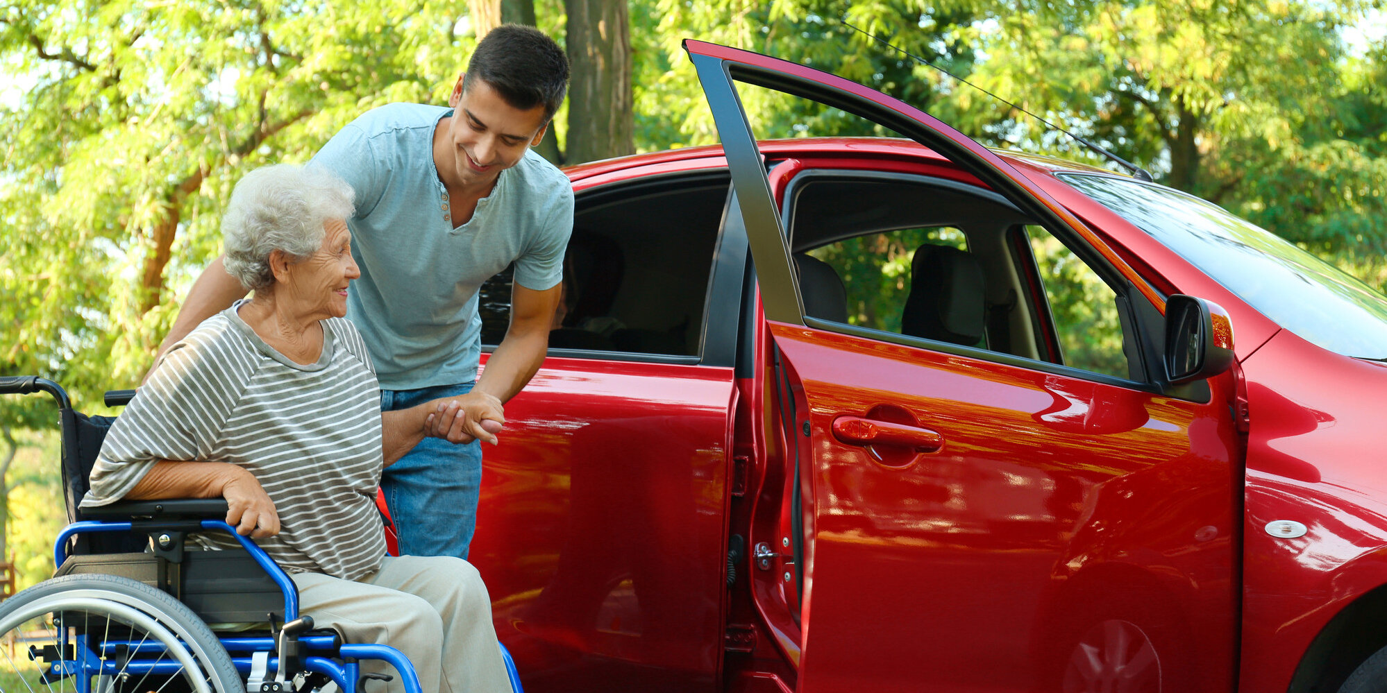 Ein junger Mann hilft einer älteren Frau aus dem Rollstuhl auf den Beifahrersitz eines roten Autos.