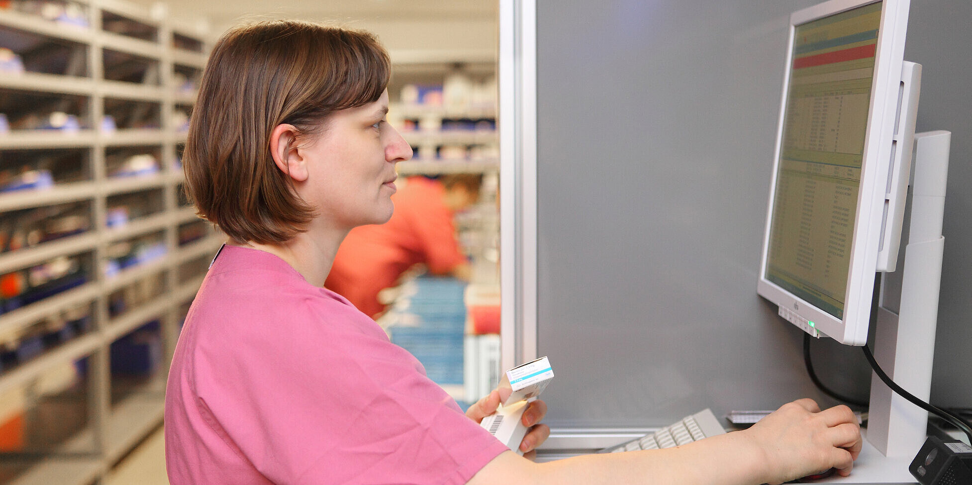 Eine Apothekerin steht vor einem Monitor und hat in der linken Hand zwei Medikamte.