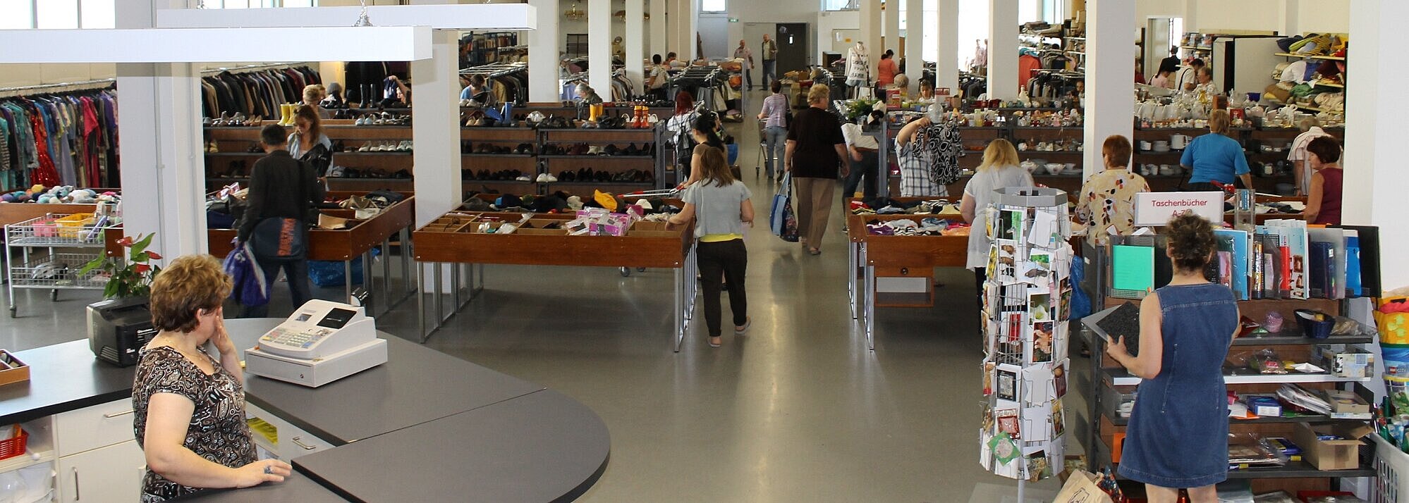 Blick in die Halle des Second-Hand-Kaufhauses Fundgrube: Viele Verkaufsregale mit diversen Angeboten wie Büchern, Kleidung und Haushaltsbedarf befinden sich links und rechts von einem langen Gang der durch die Halle führt. Viele Menschen schauen sich Artikel an. Links im Bild steht ein Tresen mit einer Kasse darauf.