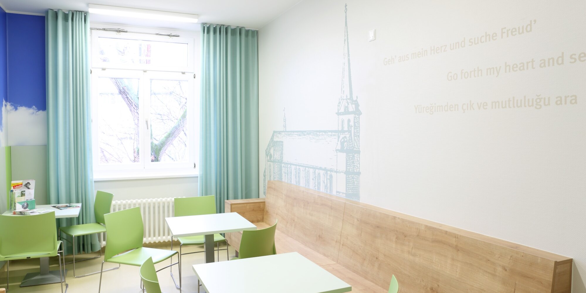 Großer und heller Aufenthaltsraum mit Sitzmöglichkeiten bestehend aus weißen Tischen und farblich passenden Stühlen. Die Wand, rechts im Raum ist zum Teil mit Holz verkleidet sowie mit einem Berlin-Motiv verziert. Das Fenster am Kopf des Raumes ist mit blauen Vorhängen versehen.