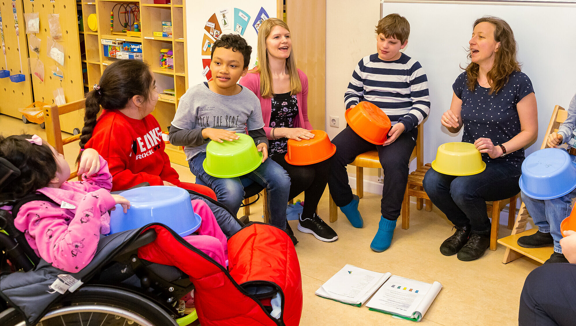 Musikunterricht: Gemeinsames Musizieren mit Alltagsgegenständen Mehrere Schüler*innen, eine Lehrerin und eine Betreuerin sitzen im Stuhlkreis. Gemeinsam singen und trommeln sie auf bunten Plastikschüsseln.