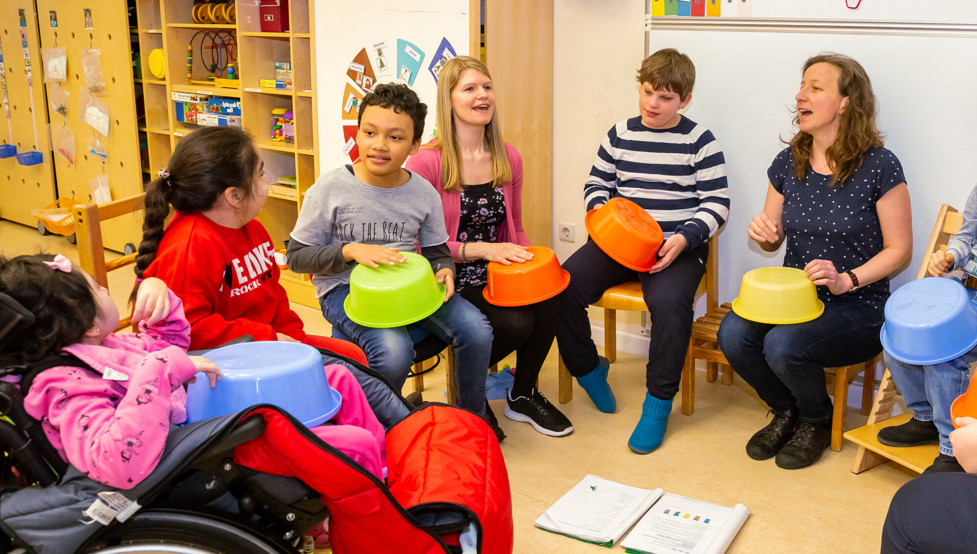 Mehrere Schüler*innen, eine Lehrerin und eine Betreuerin sitzen im Stuhlkreis. Gemeinsam singen und trommeln sie auf bunten Plastikschüsseln.