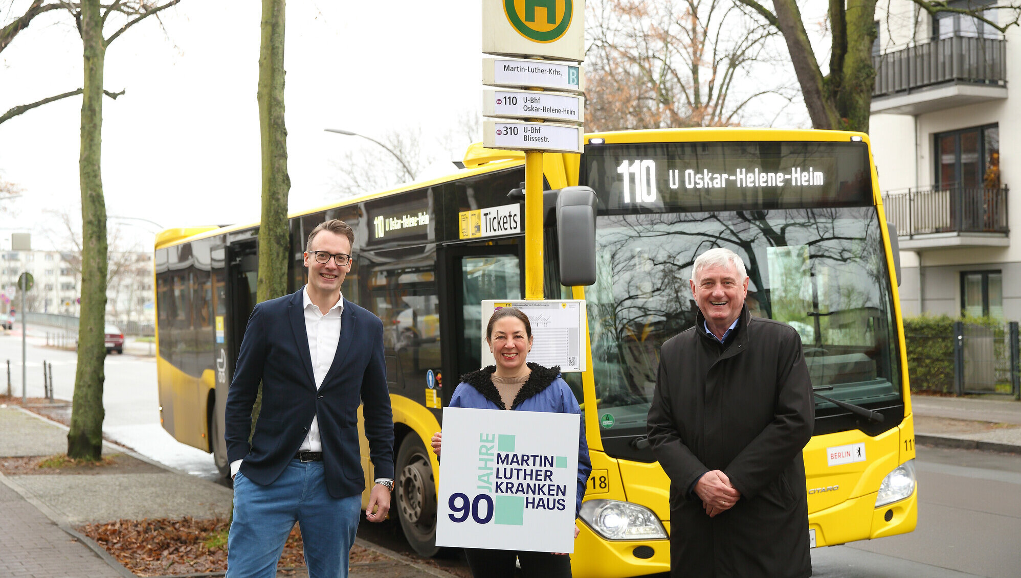 Zwei Männer und eine Frau stehen vor der Bushaltestelle „Martin-Luther-Krankenhaus“. Im Hintergrund steht ein gelber Bus.