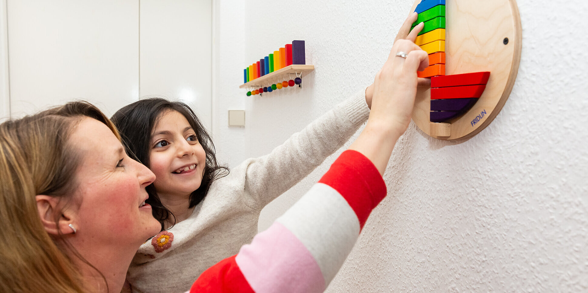 Therapien als Teil des Schulalltags Ein Schülerin und eine Betreuerin spielen mit einem bunten Wandklappspiel, um Farb- und Formempfinden spielerisch zu schulen.