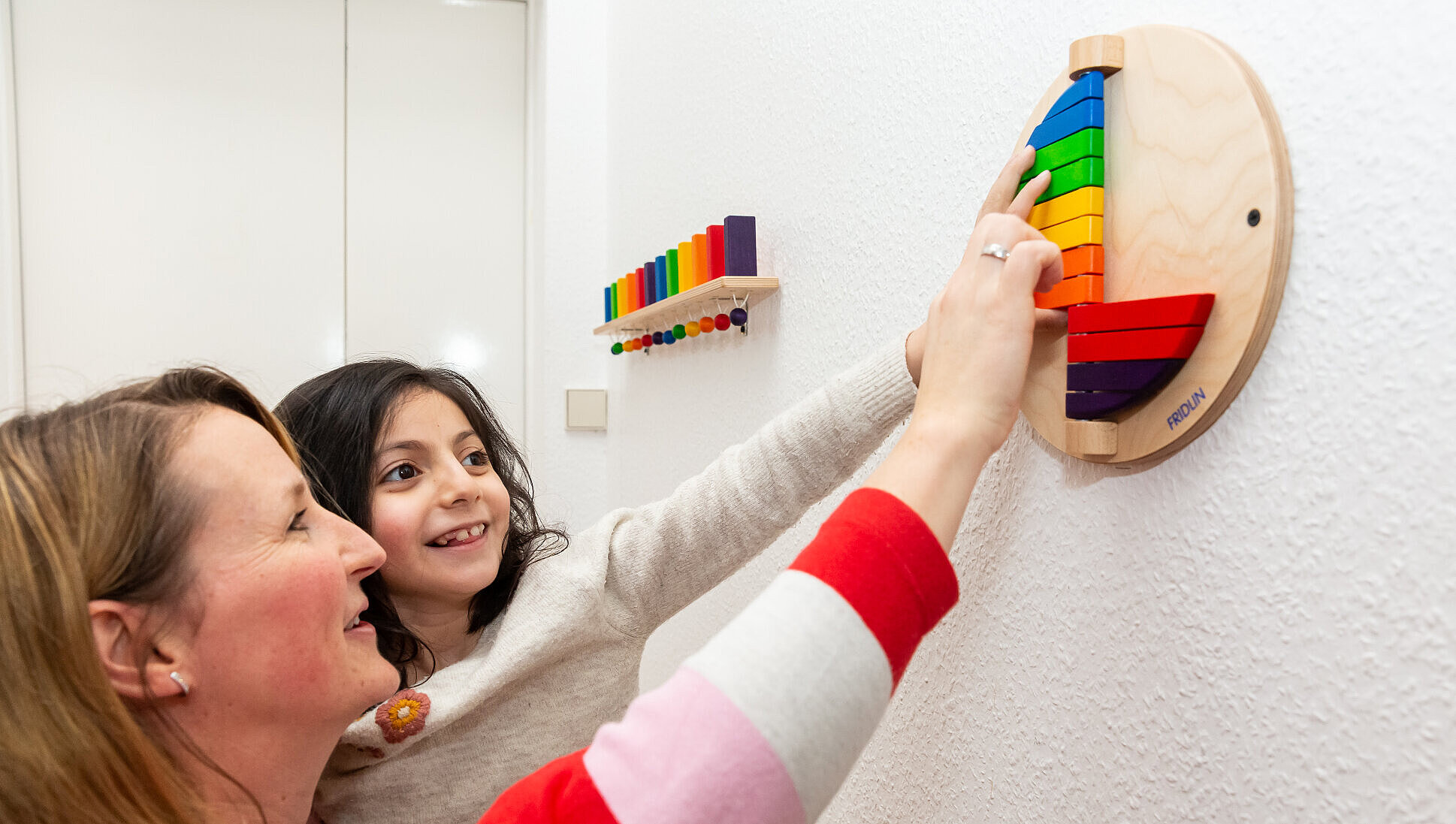 Therapien als Teil des Schulalltags Ein Schülerin und eine Betreuerin spielen mit einem bunten Wandklappspiel, um Farb- und Formempfinden spielerisch zu schulen.