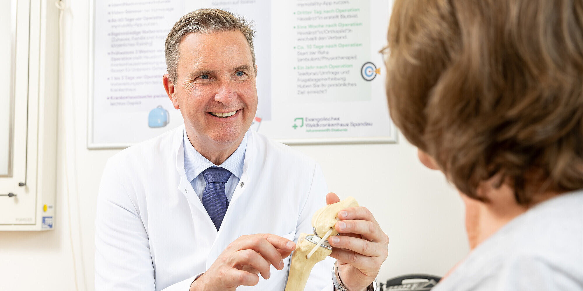 Prof. Dr. med. habil. Ulrich Nöth MHBA, Klinikdirektor der Klinik für Orthopädie und Unfallchirurgie, zeigt einer Patientin ein künstliches Kniegelenk.