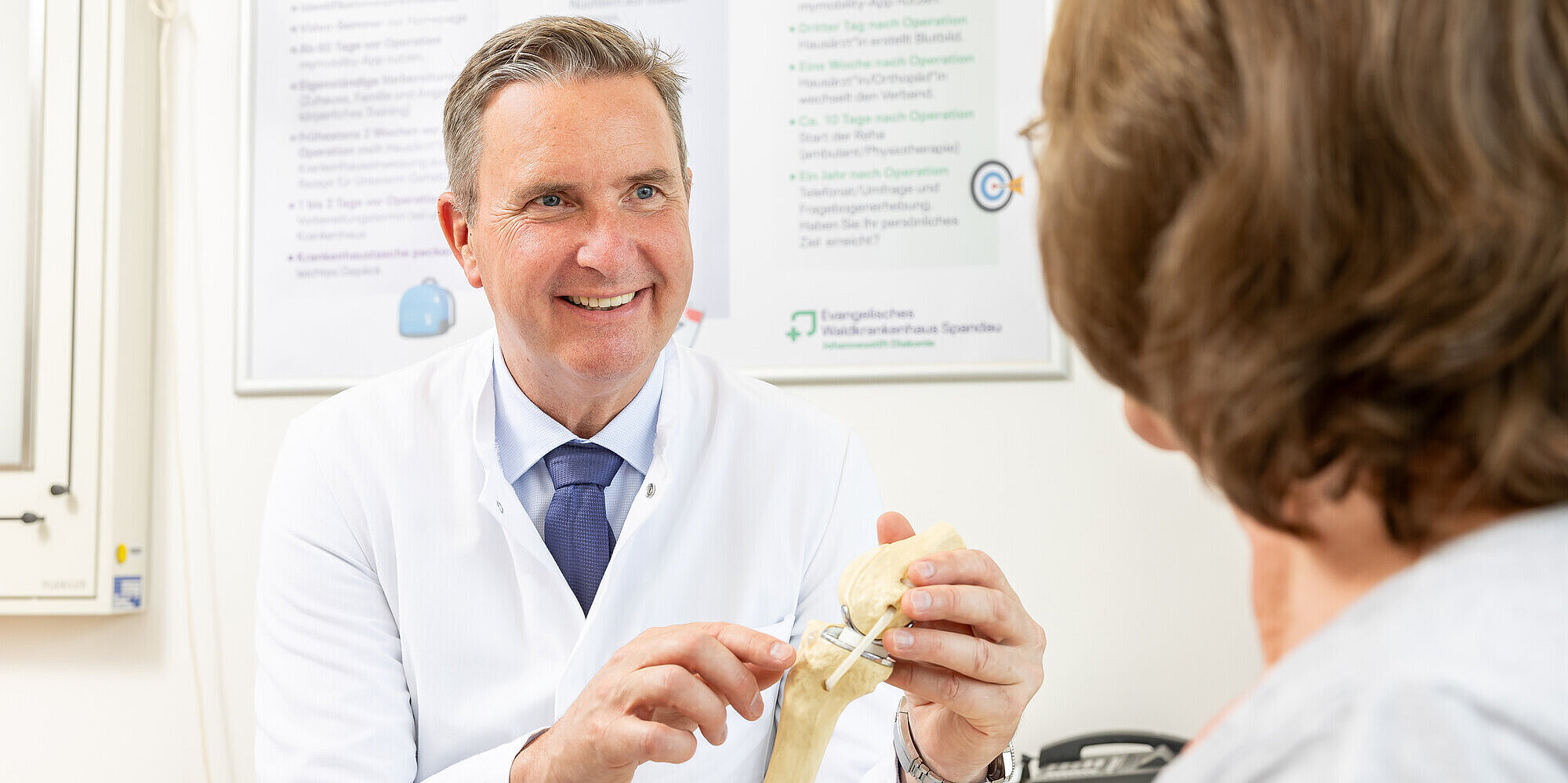 Prof. Dr. med. habil. Ulrich Nöth MHBA, Klinikdirektor der Klinik für Orthopädie und Unfallchirurgie, zeigt einer Patientin ein künstliches Kniegelenk.