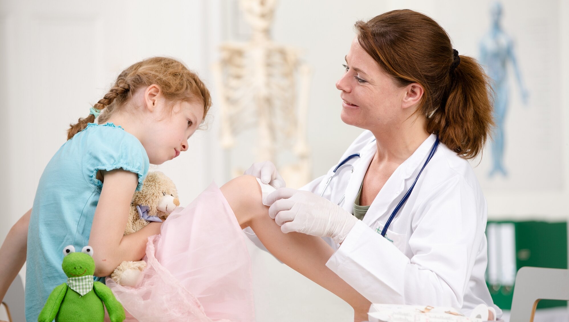 Junge Ärztin verbindet im Behandlungszimmer das Bein eines Kindes. Das Kind hat ein Kuscheltier im Arm und schaut auf das eigene Bein.