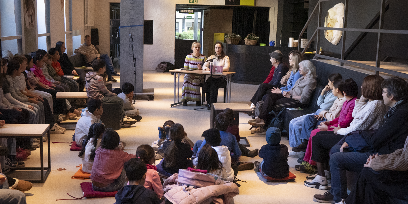 Viele Kinder in einem Raum, die zwei Vorleserinnen lauschen