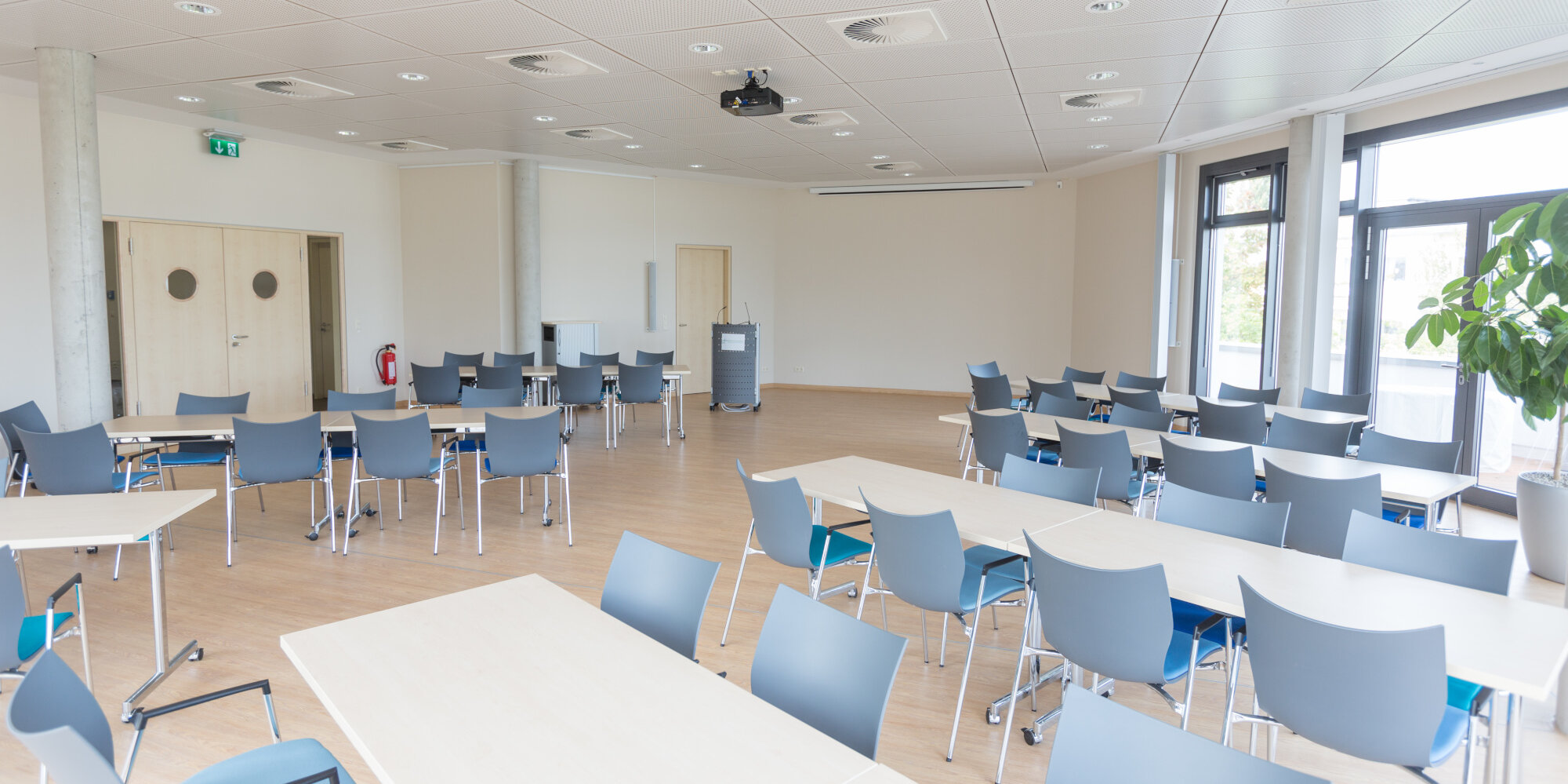 Großer und heller Konferenzraum mit Rednerpult und vielen Sitzmöglichkeiten an rechteckigen Tischen für die Teilnehmenden
