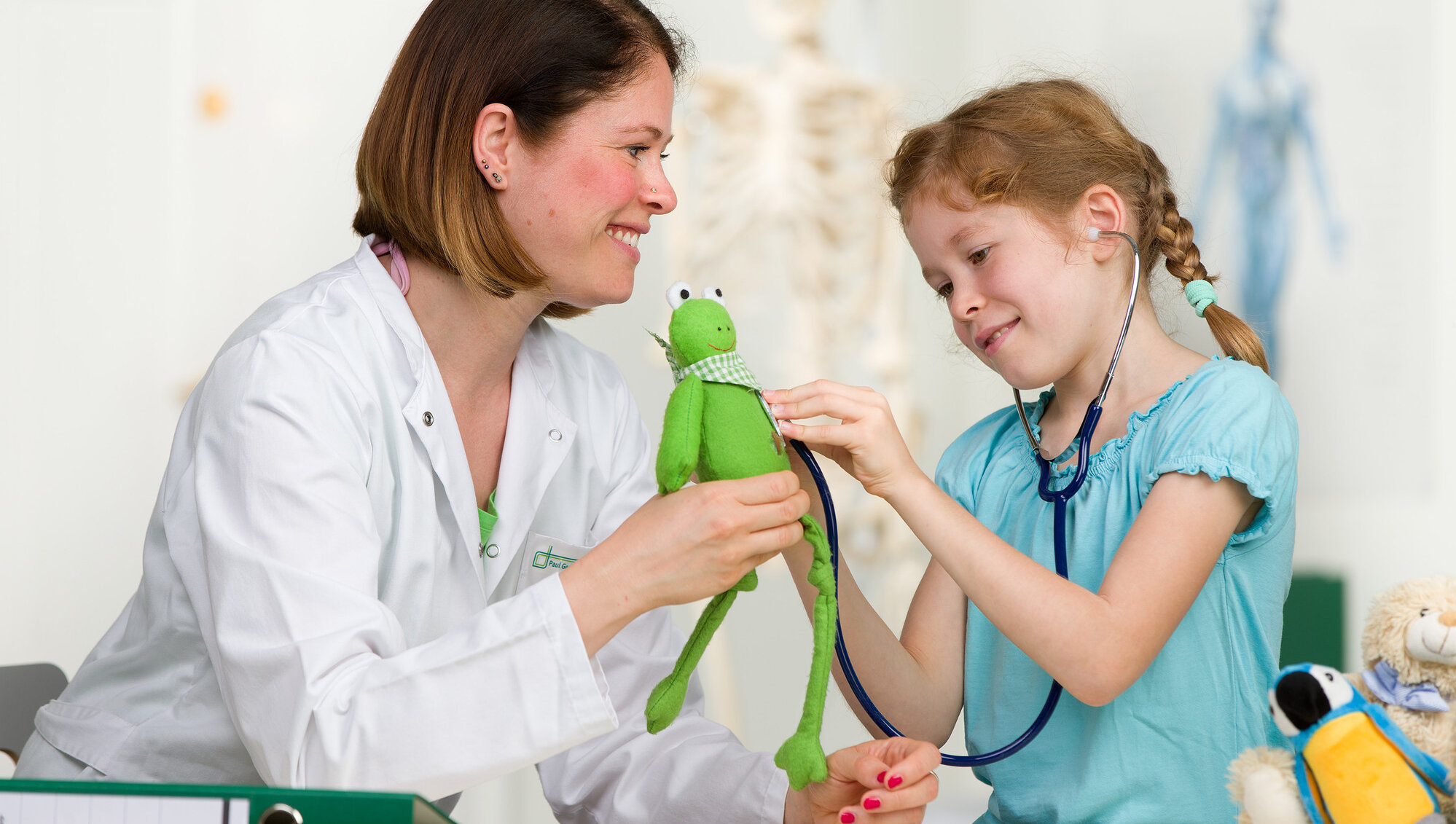 Ein Kind hört ein Kuscheltier mit dem Stethoskop ab. Eine junge Ärztin unterstützt dabei.