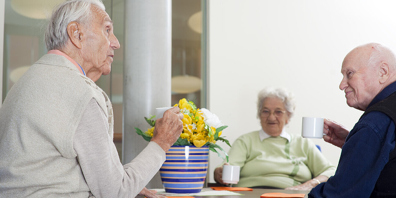 Eine Bewohnerin und zwei Bewohner sitzen am Tisch und trinken gemeinsam Kaffee.
